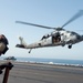 A U.S. Navy MH-60 Seahawk helicopter is hovering or taking off from the flight deck of an aircraft carrier. A flight deck crew member wearing a brown jersey and helmet with a green 'P' marking observes the helicopter from the edge of the deck. The scene appears to take place over open ocean under clear skies.