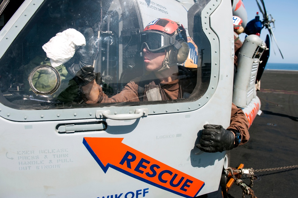 A close-up photograph of a rescue helicopter cockpit with a crew member wearing a red helmet and flight gear seated inside. A ground crew member in brown coveralls and black gloves is visible on the exterior of the aircraft. The helicopter is positioned on what appears to be a ship's flight deck, identifiable by chains securing the aircraft and ocean visible in the background.