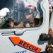 A close-up photograph of a rescue helicopter cockpit with a crew member wearing a red helmet and flight gear seated inside. A ground crew member in brown coveralls and black gloves is visible on the exterior of the aircraft. The helicopter is positioned on what appears to be a ship's flight deck, identifiable by chains securing the aircraft and ocean visible in the background.
