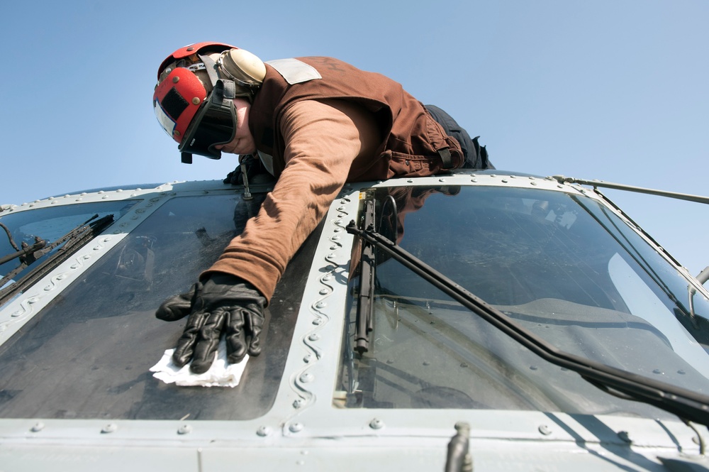 A technician or crew member wearing a red helmet with goggles and a brown flight suit is cleaning or wiping down the cockpit windshield of a military helicopter. The photo is taken from a low angle looking up, giving a dramatic perspective of the person leaning over the aircraft's nose. The clear blue sky serves as the background.