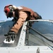 A technician or crew member wearing a red helmet with goggles and a brown flight suit is cleaning or wiping down the cockpit windshield of a military helicopter. The photo is taken from a low angle looking up, giving a dramatic perspective of the person leaning over the aircraft's nose. The clear blue sky serves as the background.