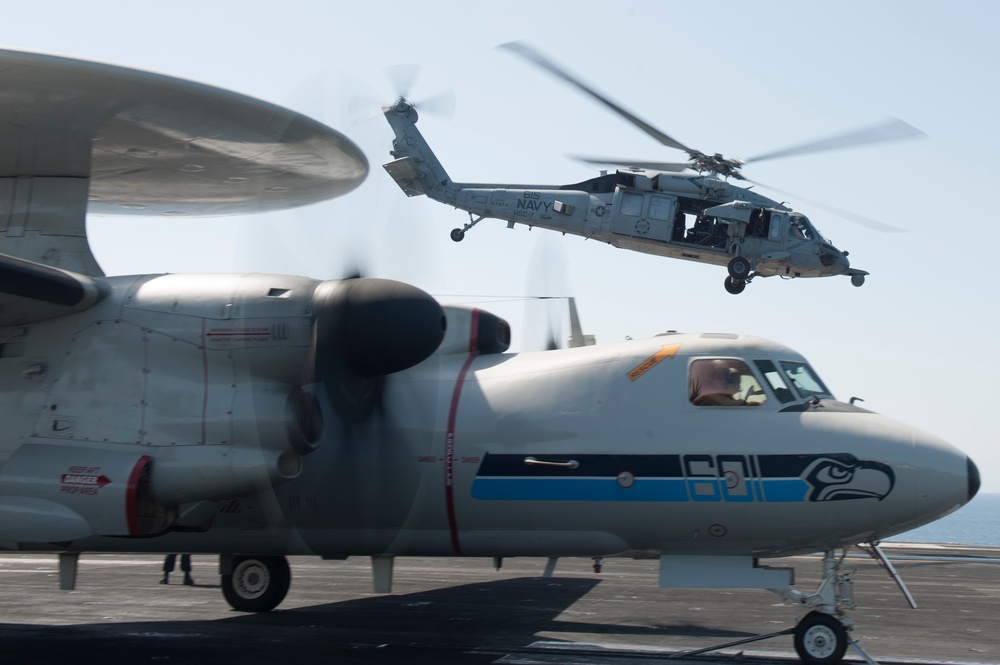The image shows a U.S. Navy aircraft carrier flight deck with two distinct aircraft visible. In the foreground is a fixed-wing turboprop aircraft (likely a C-2 Greyhound or E-2 Hawkeye) decorated with Seattle Seahawks NFL team livery including the number '601' and team logo. In the background, a Navy helicopter (MH-60 Seahawk, side number 616) is hovering or landing on the deck.
