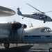 The image shows a U.S. Navy aircraft carrier flight deck with two distinct aircraft visible. In the foreground is a fixed-wing turboprop aircraft (likely a C-2 Greyhound or E-2 Hawkeye) decorated with Seattle Seahawks NFL team livery including the number '601' and team logo. In the background, a Navy helicopter (MH-60 Seahawk, side number 616) is hovering or landing on the deck.