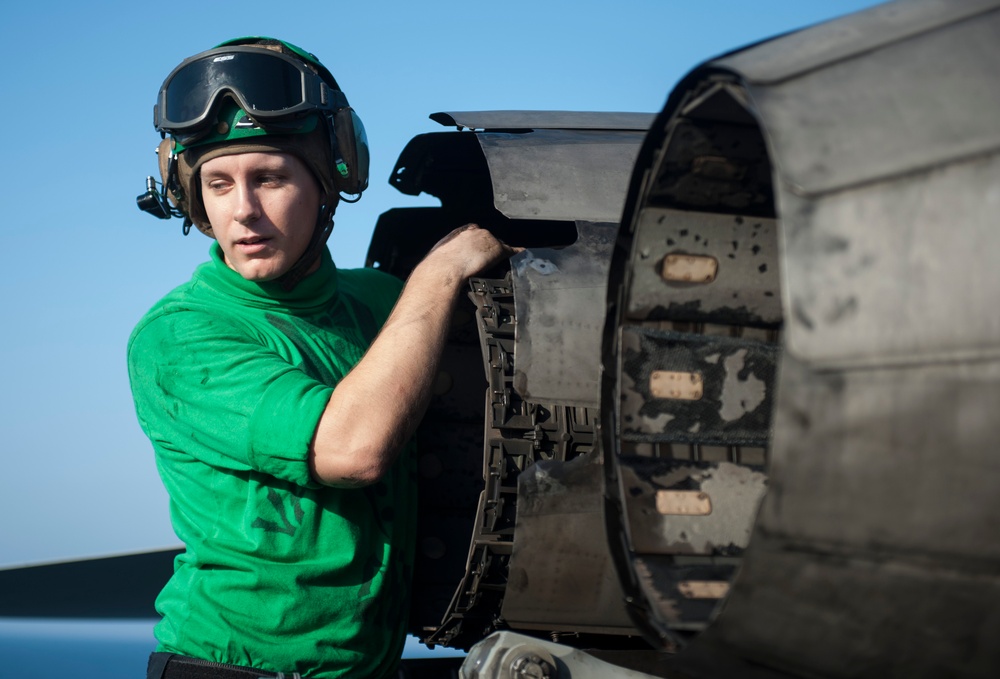 A naval aviation crew member wearing a green jersey and flight deck helmet with goggles is inspecting or working on a jet engine exhaust nozzle, likely on an aircraft carrier flight deck. The green jersey indicates the individual is likely a catapult and arresting gear crew member or aircraft handler. The engine nozzle shows visible heat damage and wear consistent with operational use.