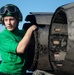 A naval aviation crew member wearing a green jersey and flight deck helmet with goggles is inspecting or working on a jet engine exhaust nozzle, likely on an aircraft carrier flight deck. The green jersey indicates the individual is likely a catapult and arresting gear crew member or aircraft handler. The engine nozzle shows visible heat damage and wear consistent with operational use.