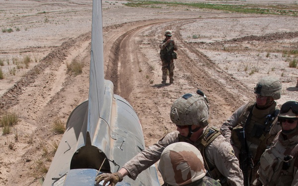 Military personnel in desert camouflage uniforms are examining or working on what appears to be a downed or grounded aircraft fuselage in an arid, desert environment. Multiple soldiers wearing helmets and tactical gear are gathered around the aircraft, with one standing guard in the background on a dirt track. The aircraft appears to be a military jet, lying on its side or at an unusual angle.