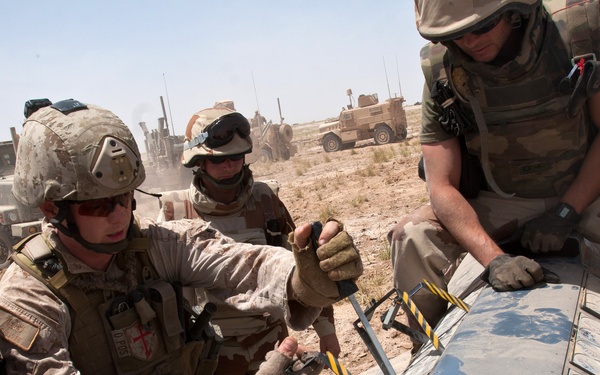 Several military personnel in desert camouflage uniforms are working together on a piece of equipment with black-and-yellow hazard markings in an arid, open environment. Military vehicles including what appear to be MRAPs or HMMWVs are visible in the background. The soldiers appear to be conducting maintenance or assembly work on a cylindrical metal component.