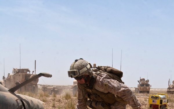 A U.S. Marine in desert MARPAT camouflage, wearing a helmet with goggles and carrying a full pack, is bent over examining wires or cables on arid ground. The scene appears to be a field operation in a desert environment, with military vehicles, what appears to be a downed or disabled helicopter, and a yellow generator visible in the background. The setting suggests an EOD (Explosive Ordnance Disposal) or recovery operation.
