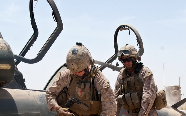 Two U.S. Marines in desert MARPAT camouflage uniforms are working near a downed or disabled dark-colored military helicopter in an arid environment. The forward Marine is handling wiring or demolition cord connected to spools on the ground, suggesting a possible explosive ordnance disposal or aircraft destruction operation. The scene appears to be a field operation in a desert combat zone.