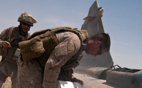 Two U.S. Marines in desert camouflage uniforms and tactical gear are working on what appears to be a downed or disabled military aircraft in a desert environment. One Marine is leaning over the aircraft fuselage and pouring a substance from a white container, producing visible vapor or smoke, possibly dry ice or a chemical agent. The setting appears to be an arid, sandy location consistent with Middle Eastern or Southwest Asian terrain.
