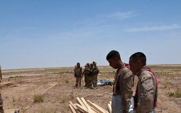 Two personnel wearing silver/aluminized proximity suits and red life vests stand in an arid, open terrain surrounded by equipment and gear. Several other uniformed individuals are visible in the background. The scene appears to be a military training exercise or emergency response drill in a desert-like environment.