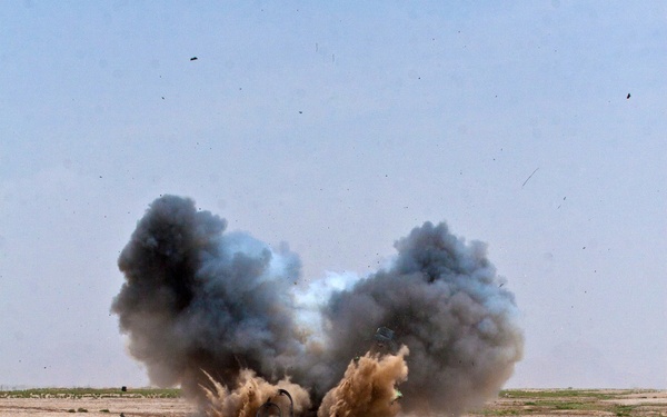 An explosion is captured mid-detonation in an arid, desert-like environment with sparse vegetation. Large plumes of dark gray smoke and tan dust billow upward from the blast site, with debris visibly scattered in multiple directions. A yellow object, possibly a container or equipment, is visible to the left of the explosion.