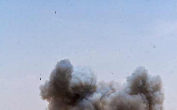 A large controlled explosion is occurring in an arid, desert-like open terrain. A significant plume of gray-brown smoke and dust rises from the blast site, with debris visibly scattered in multiple directions. A yellow piece of equipment is visible near the explosion's base, suggesting this may be a demolition or ordnance disposal operation.