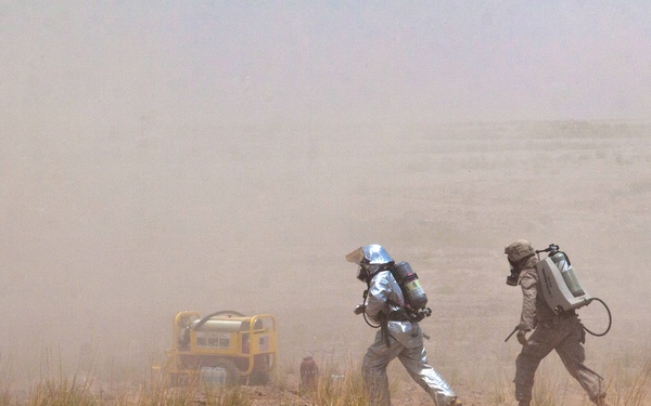 The image shows personnel in protective gear operating in a dusty, arid environment during what appears to be a CBRN (Chemical, Biological, Radiological, Nuclear) training exercise or emergency response operation. At least three individuals wearing respirators and backpack-mounted equipment are moving across dry, sparse terrain. A yellow piece of equipment or generator is visible in the background amid heavy dust or smoke.