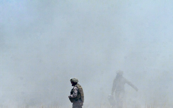 Two figures in military-style gear are moving through a heavily dust-obscured open terrain. The foreground soldier appears to be wearing body armor and a helmet, while a second figure is visible further back in the dust cloud. The ground is arid and sparse with patches of dry grass, suggesting a desert or semi-arid environment.