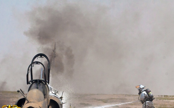 A military jet aircraft has crash-landed or been destroyed in an arid, desert-like terrain, and is actively on fire with heavy dark smoke rising into the sky. Emergency response personnel in proximity fire suits and military gear are working to extinguish the blaze using a hose and portable fire suppression equipment. The scene appears to be either an actual crash incident or a training exercise.