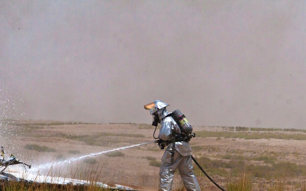 A firefighter in full proximity/aluminized protective gear and SCBA (self-contained breathing apparatus) is directing a water or foam hose stream at wreckage in an arid, desert-like environment. Smoke or dust fills the background, suggesting an active fire or post-crash scenario. A second person in military-style clothing is visible on the left near what appears to be aircraft or vehicle wreckage.