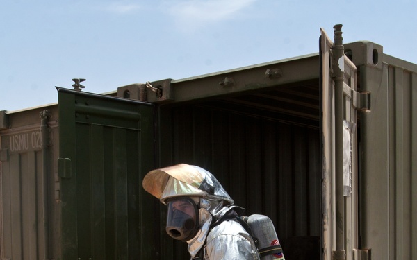 A person wearing a full aluminized proximity/hazmat suit with a Scott brand self-contained breathing apparatus (SCBA) stands at the entrance of an open military shipping container in an arid, desert-like environment. The individual appears to be conducting a hazardous materials or firefighting exercise, as indicated by the protective gear. A yellow generator or piece of equipment is visible inside the container.