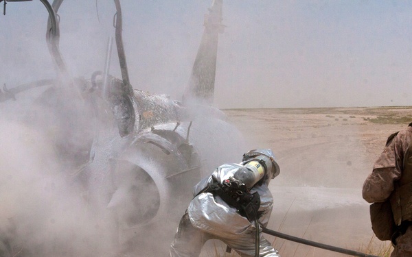 A military firefighter in a silver proximity/fire proximity suit and SCBA (self-contained breathing apparatus) is applying suppression agent to a downed or crashed aircraft in an arid desert environment. A second individual in US Marine Corps desert MARPAT camouflage uniform with tactical vest and helmet observes from the right. The scene suggests an aircraft crash response or firefighting training exercise in a desert location.