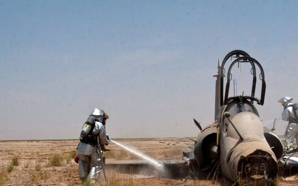 Two firefighters in full silver proximity/fire suits and SCBA (self-contained breathing apparatus) are responding to a downed military jet aircraft in an arid, desert-like terrain. The aircraft, identifiable as an F-4 Phantom based on its fuselage and canopy shape, appears to have crash-landed and shows signs of fire damage, particularly around the engine area. One firefighter is actively spraying water or suppressant foam onto the aircraft while the other approaches from the opposite side.