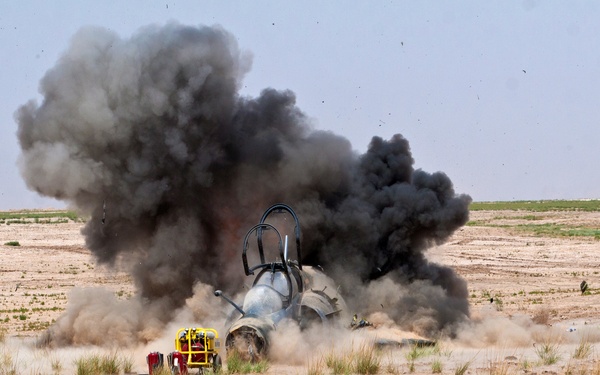 An explosion is occurring in an arid, desert-like open field, producing a large column of dark black smoke. The blast appears centered around what looks like a large cylindrical object or aircraft fuselage on the ground. Debris and dust are being thrown outward from the explosion site.