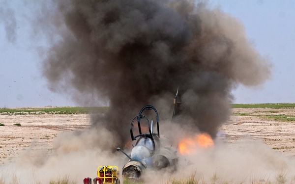 A military jet aircraft has crashed or is being destroyed on an arid, flat desert terrain. The aircraft fuselage is visible on the ground with active flames and a large column of dark smoke rising from it. A yellow piece of ground equipment is visible nearby.