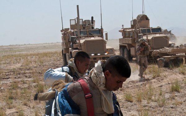 Military personnel are operating in an arid, desert environment alongside armored vehicles. A soldier in the foreground is wearing a distinctive silver/reflective protective suit and carrying heavy equipment and gear. Additional personnel and MRAP-type armored vehicles are visible in the background.