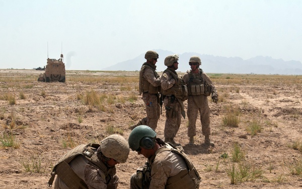 Military personnel in desert camouflage uniforms are engaged in an operation in an arid, open terrain. Two soldiers in the foreground are crouched down working with a heavy chain, while three others stand conversing in the middle ground. Additional military vehicles and personnel are visible in the background against a mountainous horizon.