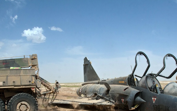 A damaged or wrecked military jet aircraft is being dragged or recovered by a heavy military truck using chains in an arid desert environment. The aircraft appears to be a French Dassault Mirage fighter jet, lying on its side or belly on sandy ground. A second military vehicle is visible on the right side of the frame.