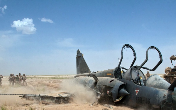 A damaged or destroyed military jet aircraft lies on its side in a desert environment, being dragged or moved by a chain attached to a military vehicle. Personnel in military uniforms are visible in the background observing the scene. A military armored vehicle is visible to the right of the downed aircraft.