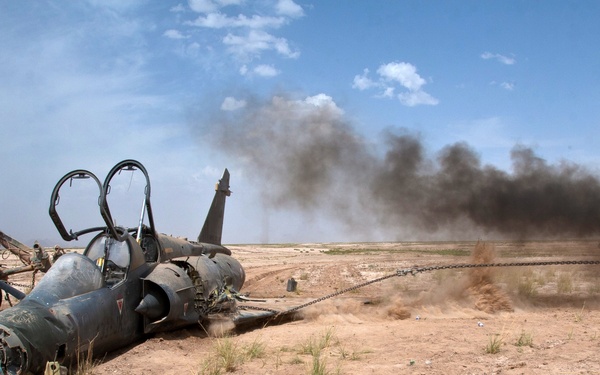 A damaged or decommissioned military jet aircraft is lying on its side in an arid desert environment. Heavy military vehicles are connected to the aircraft via chains, appearing to drag or demolish it. Black smoke is rising in the background, suggesting burning or destruction activity nearby.