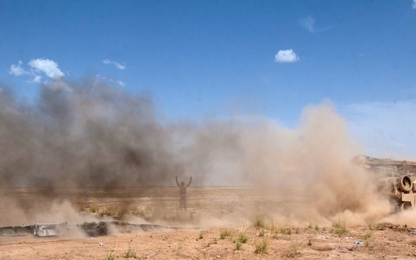 A desert combat or training scene showing a tan/sand-colored armored military vehicle generating large dust and smoke clouds in an arid environment. A human figure with arms raised is visible in the background amid the dust cloud. The scene appears to depict active military operations in a desert terrain.
