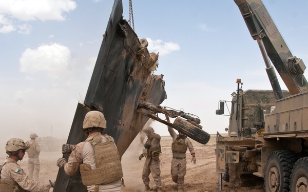Military personnel in desert camouflage uniforms are conducting a recovery or salvage operation in an arid, dusty environment. A large military crane/recovery vehicle is lifting heavily damaged and twisted metal debris, likely wreckage from a destroyed vehicle. Several soldiers are assisting on the ground, guiding and stabilizing the debris during the lift.