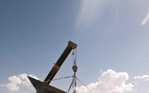 A military crane vehicle is lifting a large, heavily damaged aircraft component — appearing to be a wing or fuselage section — in an arid desert environment. The wreckage bears a visible roundel insignia with red, white, and blue concentric circles, consistent with French military aviation markings. Personnel in military attire are visible nearby, and a second vehicle is visible in the background raising dust.