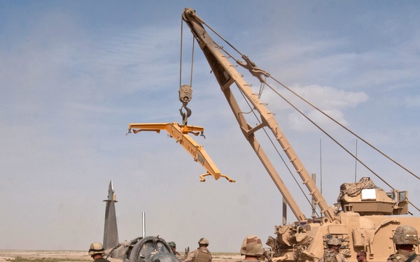 Military personnel are engaged in a recovery operation in an arid desert environment. A large crane vehicle is being used to lift or reposition what appears to be a downed or damaged aircraft fuselage. Multiple soldiers in combat gear and desert camouflage are supervising and assisting the operation.