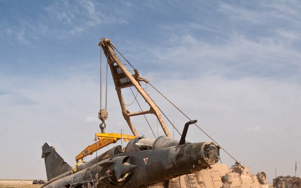 A damaged military jet aircraft is being recovered using a large yellow crane mounted on an armored recovery vehicle in an arid desert environment. Several military personnel in camouflage uniforms are observing and assisting the recovery operation. The aircraft appears heavily damaged, with its nose section destroyed and fuselage showing significant structural damage.