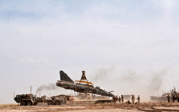 A military jet aircraft, appearing to be a Dassault Mirage-type fighter, is being transported on a heavy-duty flatbed trailer in an arid desert environment. Multiple military vehicles and personnel are present, with smoke or dust visible in the background. The operation appears to be a recovery or relocation effort of the aircraft in what looks like a Middle Eastern or Central Asian desert setting.