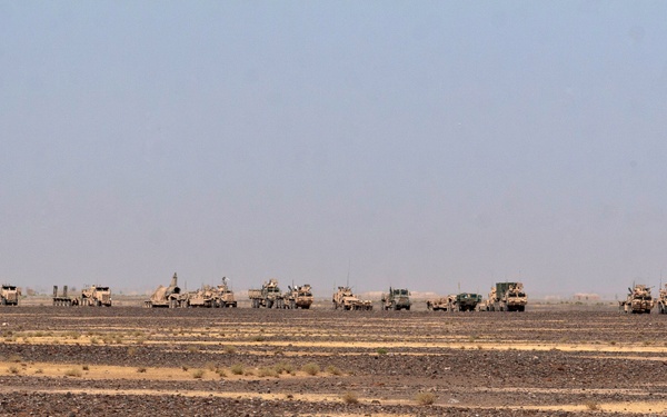 A large convoy of military vehicles is staged or moving across an arid desert terrain. The vehicles appear to be tan/sand-colored, consistent with desert camouflage paint schemes. The convoy spans a wide horizontal line across the mid-ground of the image against a hazy sky.