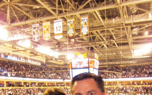 Two people, a young woman and an older man, pose for a photo together at what appears to be an indoor hockey arena filled with spectators. The arena is brightly lit with a visible ice rink in the background, championship banners hanging from the rafters, and a large scoreboard/jumbotron overhead. A slogan reading 'FUELLED BY PASSION' is visible on the arena signage.