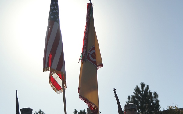 A military color guard unit stands at attention on a paved surface, holding the United States flag and what appears to be a unit guidon or organizational flag. The soldiers are dressed in US Army ACU (Army Combat Uniform) digital camouflage pattern. The image is backlit by bright sunlight, creating strong silhouette effects on the figures.
