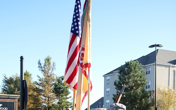 Four U.S. Army soldiers in ACU (Army Combat Uniform) digital camouflage are standing in a color guard formation on a sidewalk or roadway. Two soldiers are carrying flags including the American flag and what appears to be a unit guidon, while two others carry rifles. The setting appears to be a military installation or civilian area with buildings and trees visible in the background.