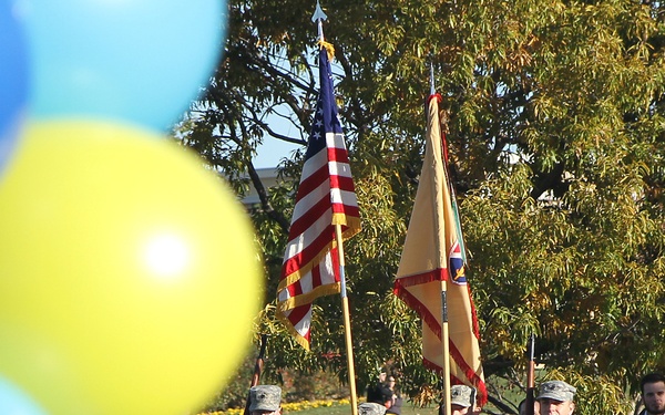 A military color guard in U.S. Army ACU (Army Combat Uniform) camouflage is marching in formation carrying the U.S. national flag and what appears to be a unit guidon or organizational flag. The ceremony takes place outdoors on a paved path surrounded by trees with autumn foliage. Blue and yellow balloons in the foreground suggest a celebratory or commemorative public event.