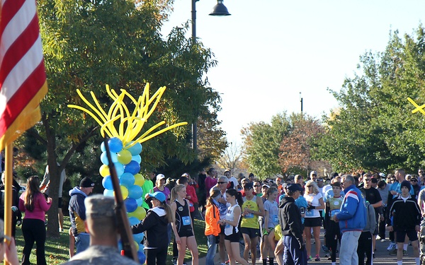 An outdoor running race or fun run event is taking place on a paved road or path, with participants gathered near a decorated start or finish line area. Blue and yellow balloon columns with starburst toppers serve as decorative archways marking the course. Military personnel in camouflage uniforms carrying flags, including an American flag, are present at the foreground, suggesting a military-affiliated or patriotic event.