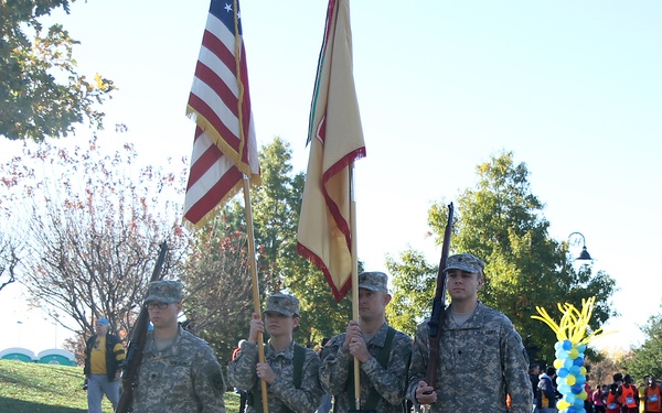 Four military personnel in U.S. Army ACU (Army Combat Uniform) camouflage are performing a color guard ceremony on a paved road. Two members carry flags — the U.S. national flag and what appears to be a unit or state guidon — while two others carry rifles at their sides. In the background, a crowd of participants and spectators is gathered near blue and yellow balloon decorations, suggesting a public event such as a race or community gathering.