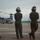 Three military personnel in olive drab flight suits and flight helmets stand on a tarmac observing a tiltrotor aircraft, identified as a V-22 Osprey, on the flight line. The individuals are viewed from behind, with helmet markings partially visible in red and green text. The scene appears to be a flight operations environment under overcast skies.