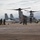 A U.S. Marines MV-22 Osprey tiltrotor aircraft is parked on a tarmac apron, surrounded by military personnel in flight suits and camouflage uniforms. The aircraft's rotors are in the horizontal (airplane mode) position with red protective covers on the engine nacelles. Personnel appear to be conducting pre-flight or post-flight checks and briefings.