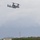 A military tiltrotor aircraft, identifiable as a Bell Boeing V-22 Osprey, is captured in flight over what appears to be a military airfield or base. The aircraft is in a transitional flight configuration with rotors tilted, flying at low altitude against an overcast sky. Ground infrastructure including power transmission towers, a low building, and a paved runway strip are visible in the background.
