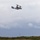 A military tiltrotor aircraft, identifiable as a Bell Boeing V-22 Osprey, is captured in low-altitude flight against an overcast sky. The aircraft appears to be in airplane mode with its rotors tilted forward for horizontal flight. The scene is set over a coastal or rural landscape with visible vegetation and power transmission infrastructure in the background.