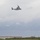 A military tiltrotor aircraft, identifiable as a Bell Boeing V-22 Osprey, is captured in flight against an overcast sky. The aircraft appears to be in airplane mode with rotors tilted upward, flying at moderate altitude. In the background, a multi-story building and power transmission towers are visible above a treeline.