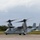 A U.S. Marines MV-22 Osprey tiltrotor aircraft is on a runway with its rotors spinning, indicating it is either preparing for takeoff or has just landed. The aircraft bears the number '04' and 'MARINES' markings on its fuselage. A second aircraft tail section is visible in the background, and the setting appears to be a military airfield surrounded by tropical vegetation.